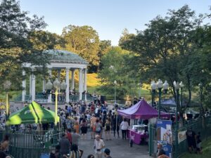 A crowd gathers at the Roger Williams Park gazebo for an outdoor event. Tents from RWP Conservancy and vendors are set up nearby.