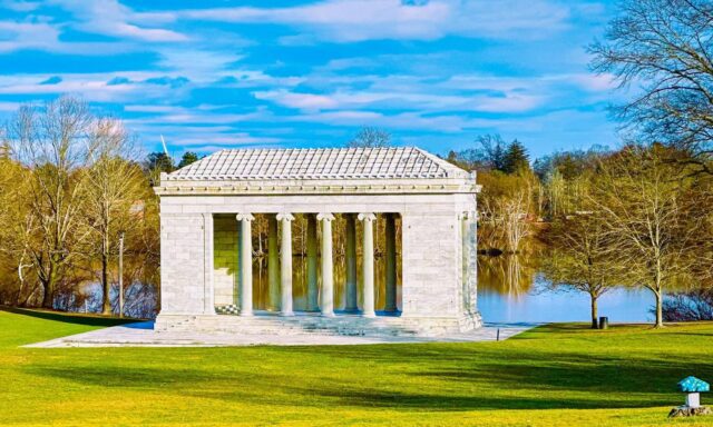 A marble Greek-style pavilion with columns stands by a serene lake, surrounded by trees and a vibrant green lawn under a blue sky.