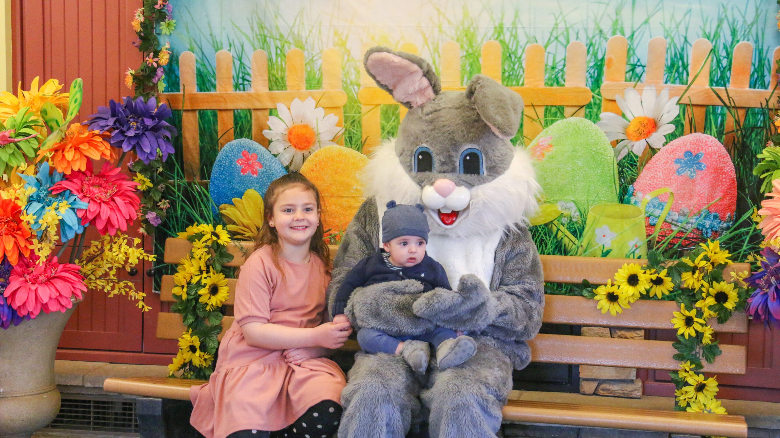 A girl and a baby sit with the Easter Bunny on a bench, surrounded by colorful flowers and a festive backdrop with decorated eggs.