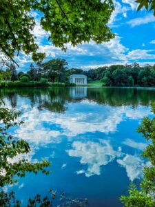 A white temple with columns is reflected in a serene lake, framed by lush green trees and a vibrant blue sky with clouds.