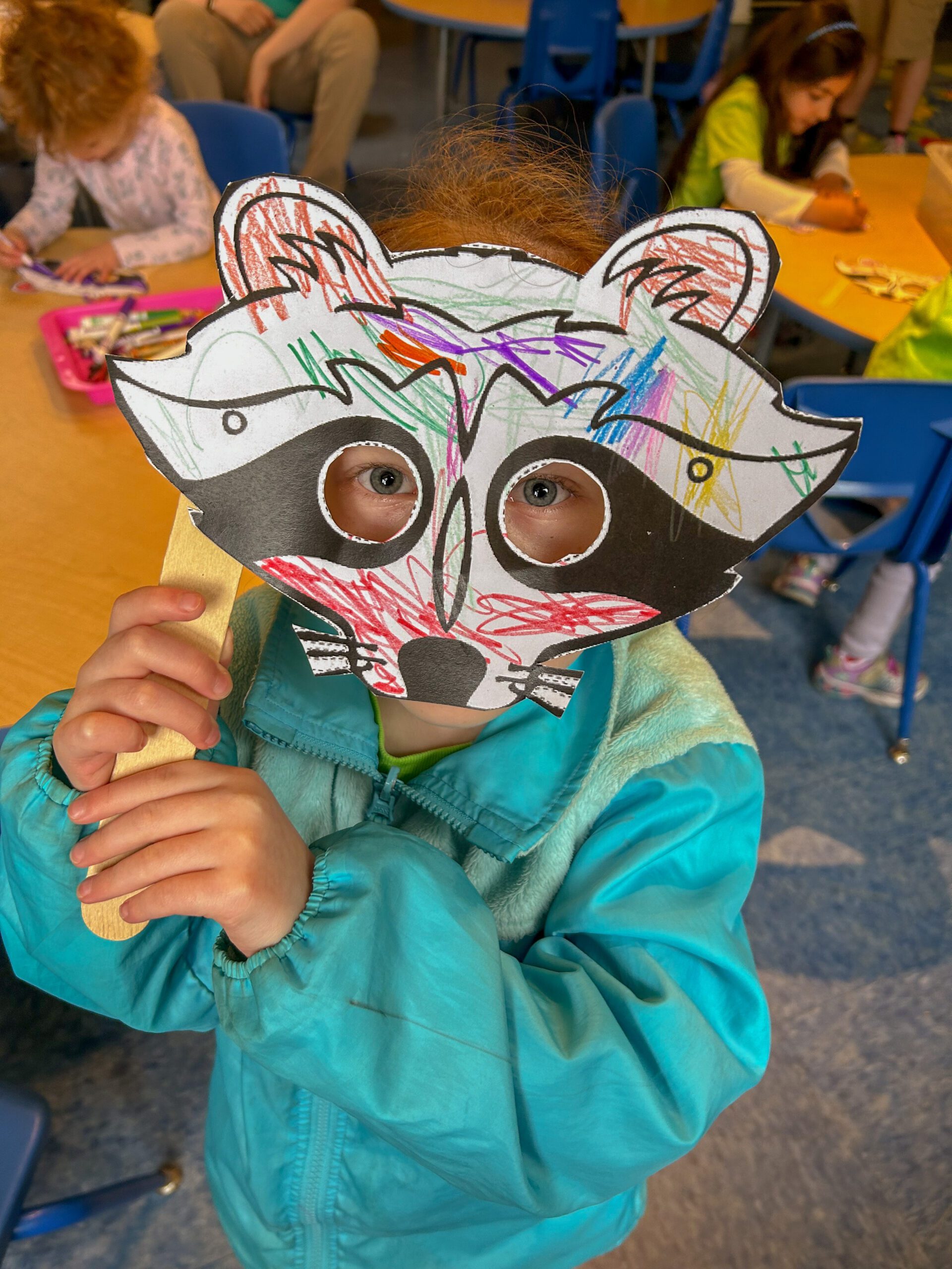 A child in a blue jacket holds a colorful raccoon mask at Zoo-Cation Day Camp, with other children crafting in the background.