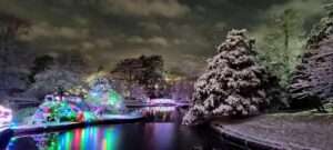 A snowy landscape at night with colorful lights illuminating trees and a bridge, reflecting on the water below.