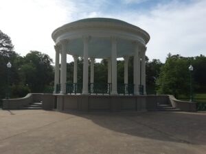 A white, circular bandstand with tall columns and a blue-green roof stands in a park setting, surrounded by trees and lampposts.