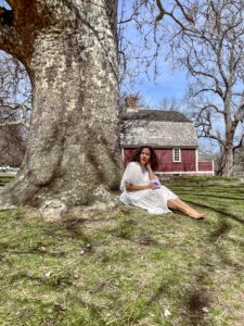 A woman in a white dress sits under a large tree, writing in a notebook. Behind her is a red house with a shingled roof.