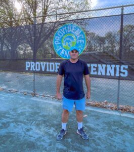 A man in a blue cap and athletic wear stands on a tennis court in front of a "Providence Tennis" sign and logo. Trees are in the background.
