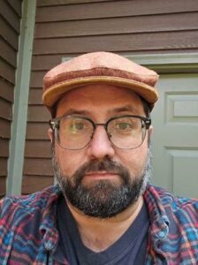 A man with glasses and a brown cap stands in front of a wooden wall and door.