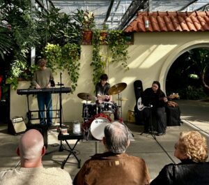 Three musicians perform in a lush greenhouse, with an audience seated in front.
