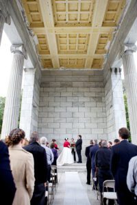 A couple exchanges vows under a grand structure, surrounded by seated guests.