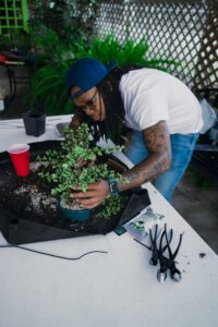 A person with long hair plants a small tree in a pot, surrounded by gardening tools and a red cup.