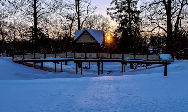 A wooden bridge leads to a small cabin at sunset, surrounded by snow-covered ground and trees.