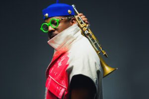 A man with a trumpet rests on his shoulder, wearing a colorful outfit and round green glasses.