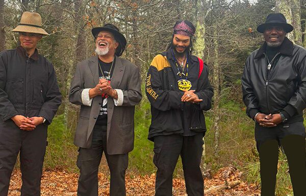 Four men stand together outdoors, smiling and clasping their hands, surrounded by trees.