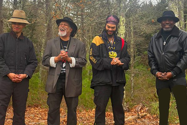 Four men stand together outdoors, smiling and clasping their hands, surrounded by trees.