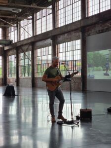 A musician plays guitar and sings on stage in a spacious venue with large windows.
