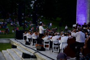 An orchestra performs on stage, with a conductor leading and an audience seated on the grass.
