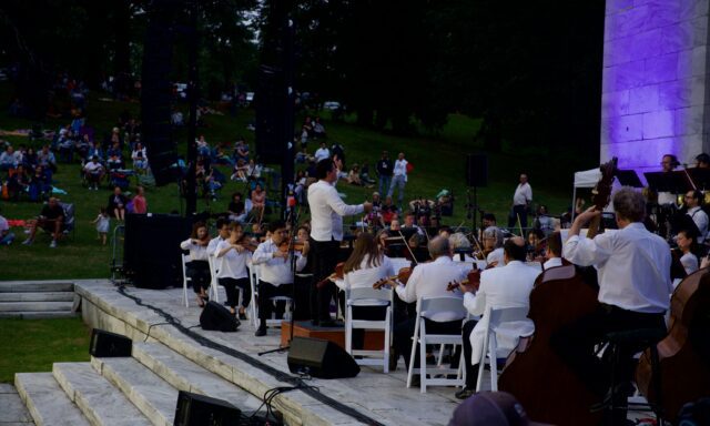 An orchestra performs on stage, with a conductor leading and an audience seated on the grass.