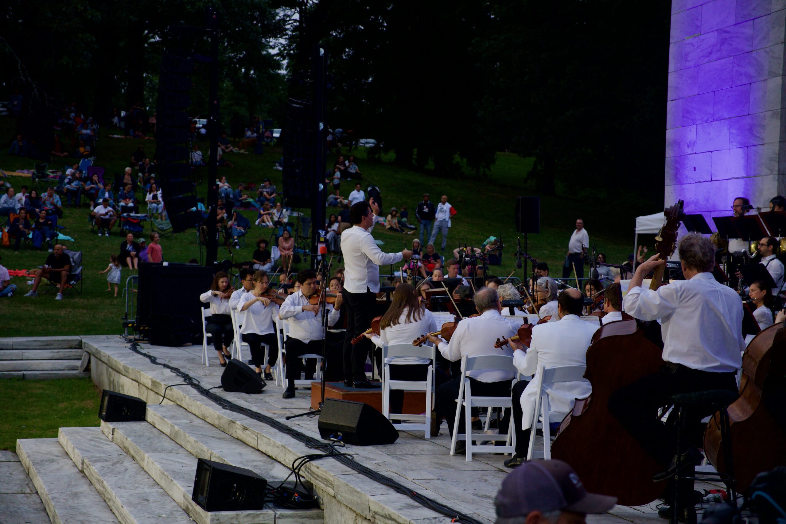 An orchestra performs on stage, with a conductor leading and an audience seated on the grass.